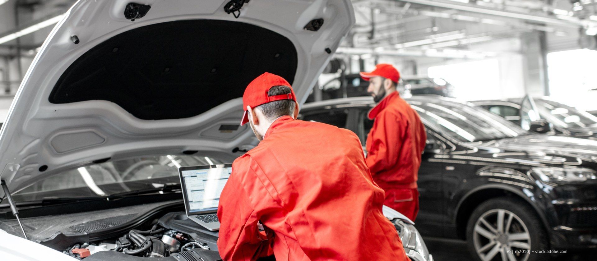 Mechaniker in roten Arbeitskleidung untersuchen in einer Autowerkstatt einen schwarzen Wagen mit geöffneter Motorhaube.