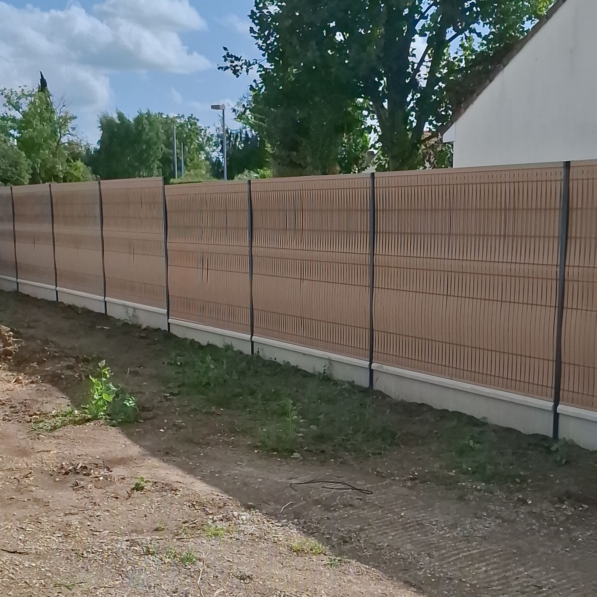 Clôture beige avec poteaux noirs, donnant sur une cour gazonnée et un bâtiment.