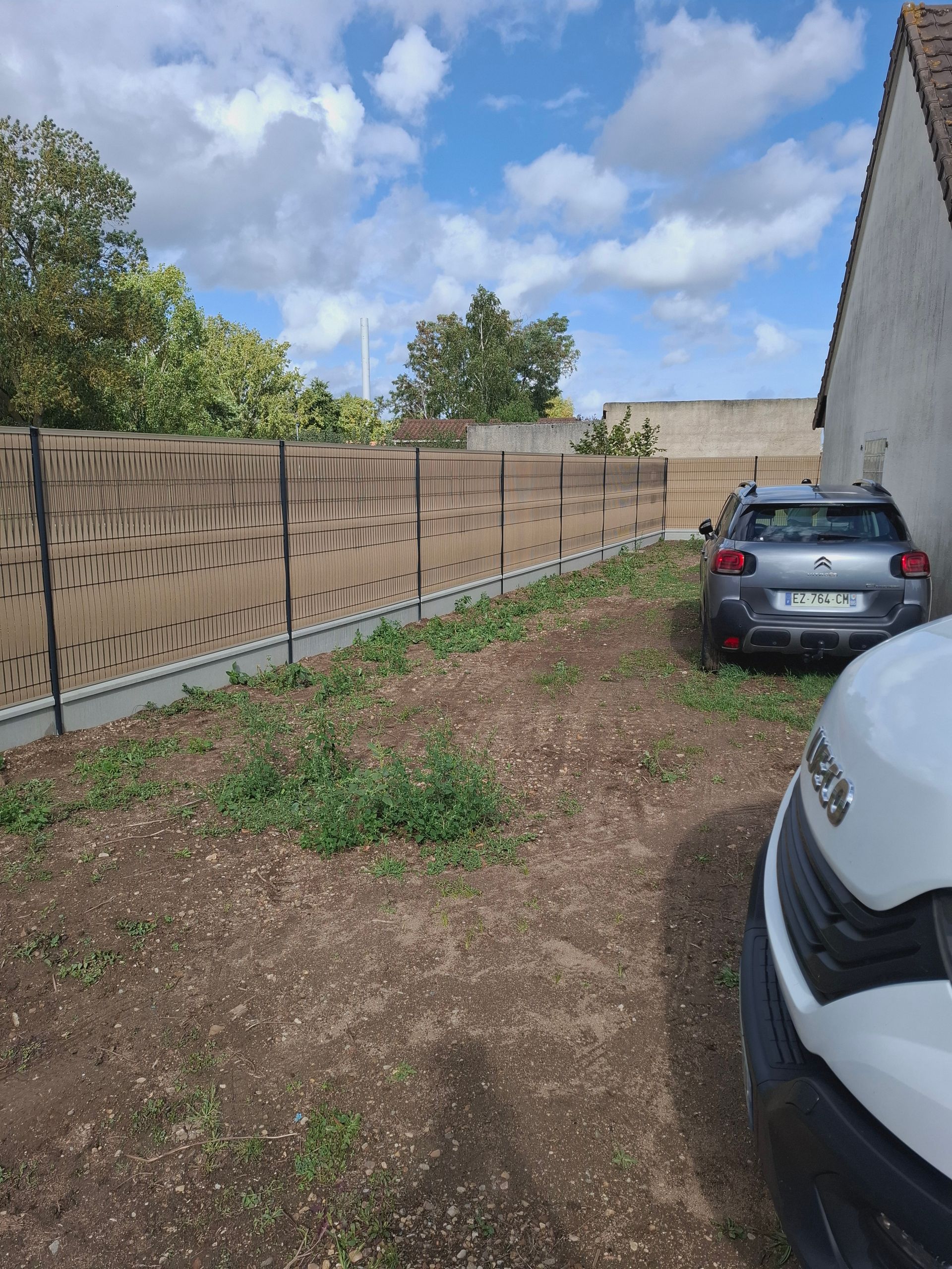 Une cour clôturée avec une voiture garée à côté d'un bâtiment sous un ciel bleu.