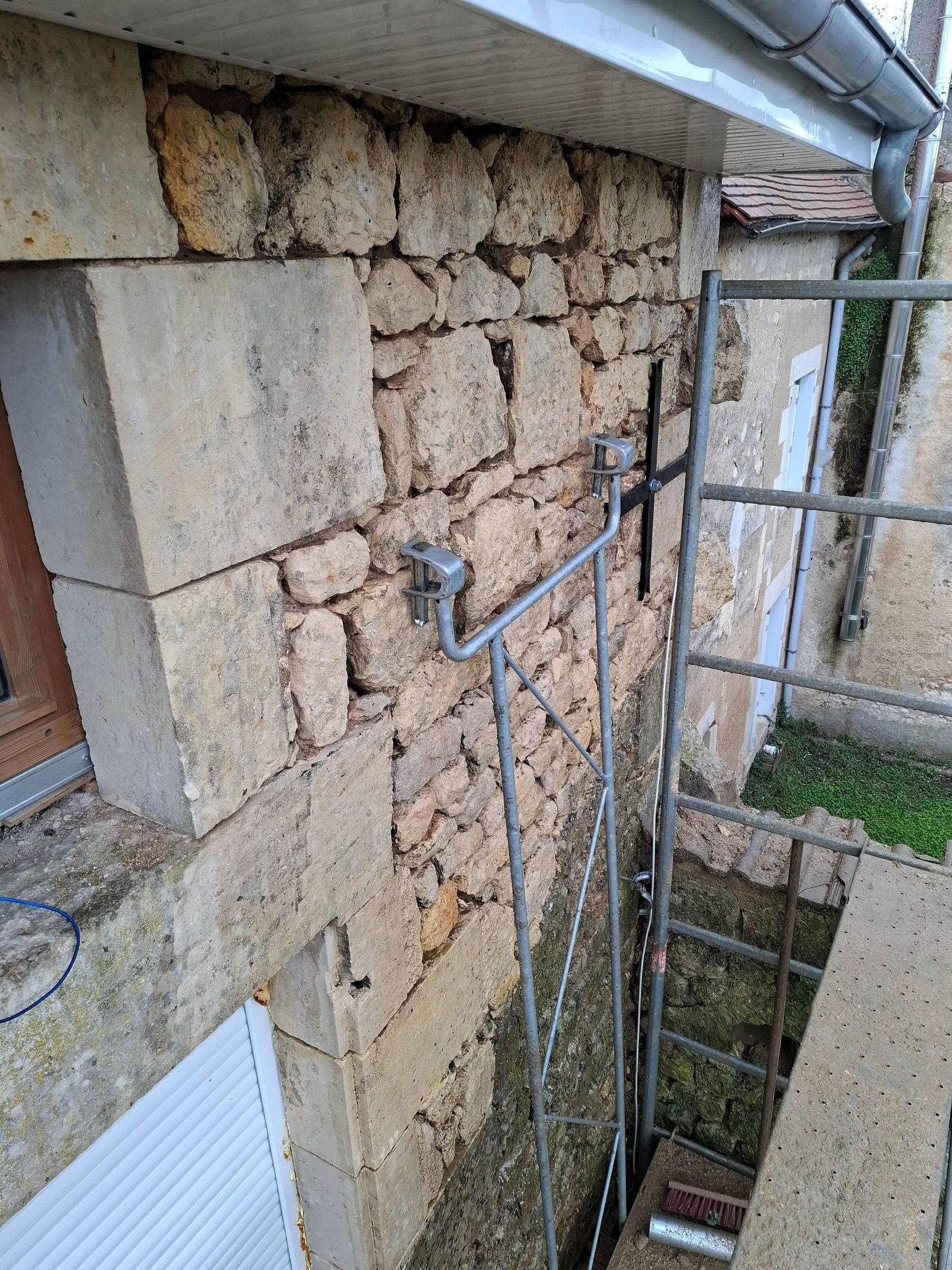 Mur en pierre avec une échelle métallique fixée, près d'un caniveau et du bord d'un bâtiment.