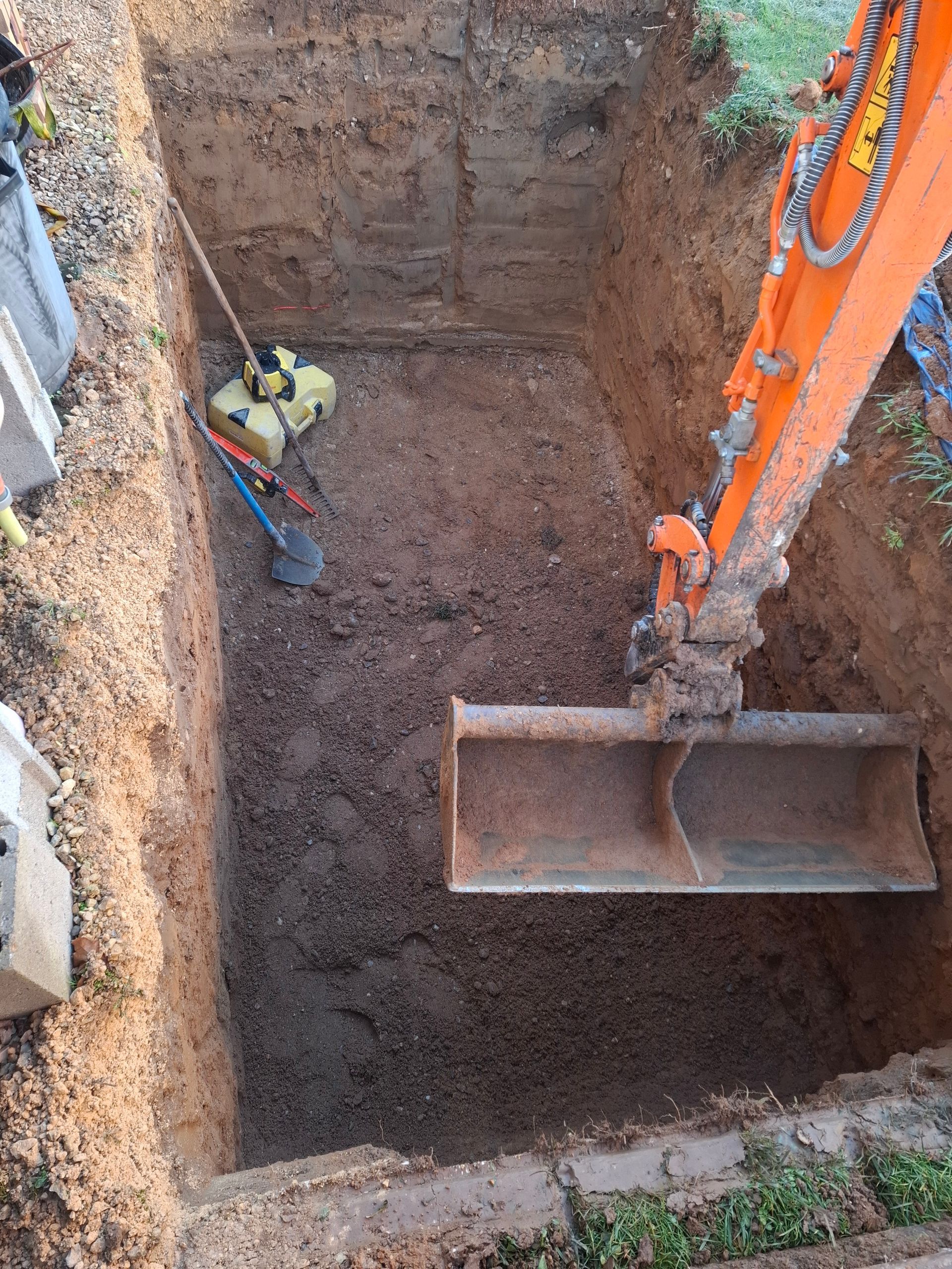 Godet d'excavatrice dans une fosse rectangulaire ; un compacteur est visible à l'intérieur.