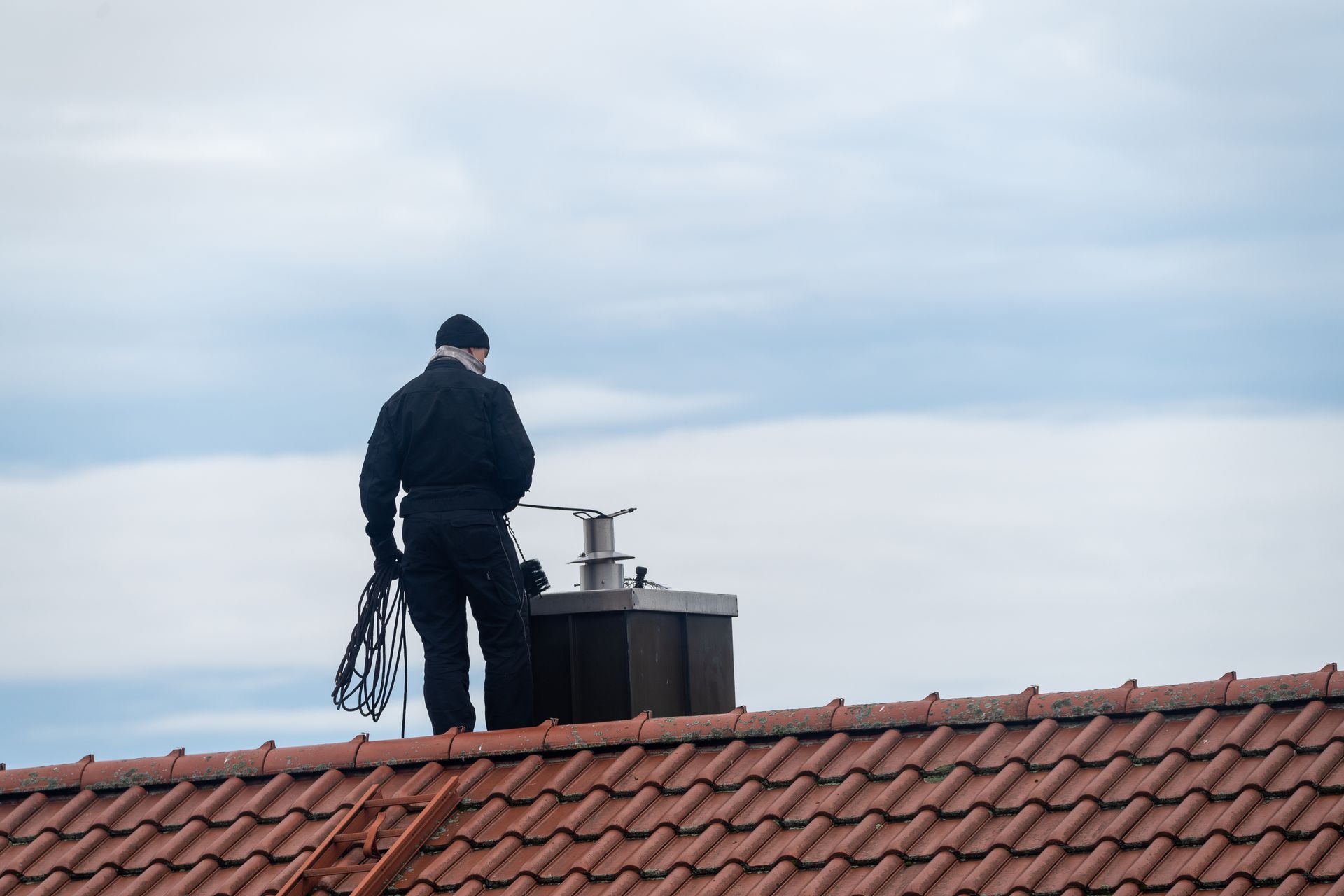 Un ramoneur sur un toit près d'une cheminée, vêtue de vêtements sombres, ciel nuageux.