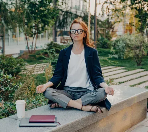 Una mujer pelirroja con gafas medita al aire libre en un banco. Está en posición de loto con los ojos cerrados en un parque.