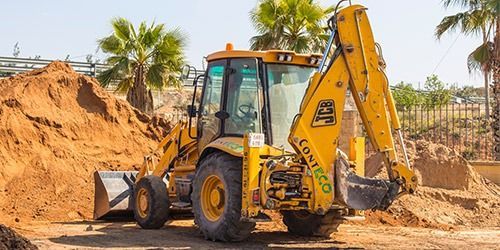 Retroexcavadora JCB amarilla en una obra, recogiendo tierra. Palmeras al fondo, día soleado.