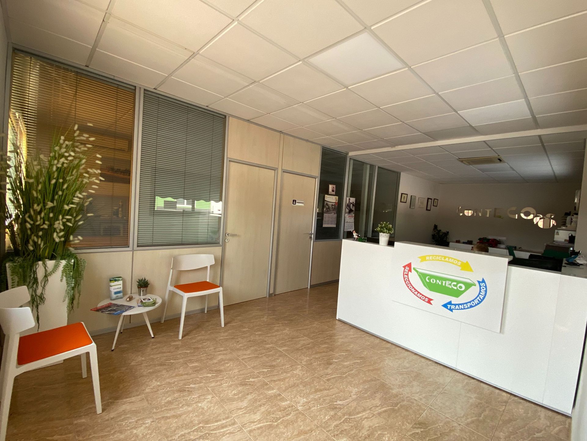 Área de recepción de oficina con escritorio blanco, sillas y planta en maceta.