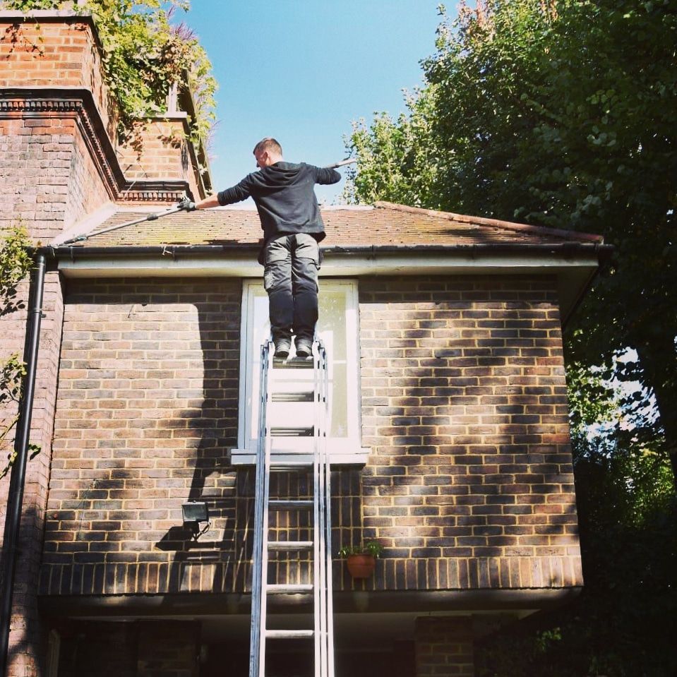 Person standing on a ladder reaching toward a brick roof in a sunny backyard