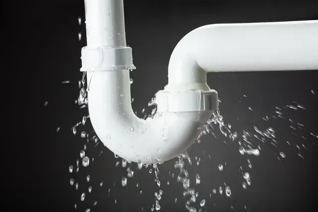 Codo de tubería blanca con fugas que rocía agua sobre un fondo oscuro.