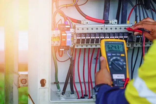 Un electricista prueba un panel de circuito con un multímetro; los cables son visibles. La lectura es de 624,3 voltios.