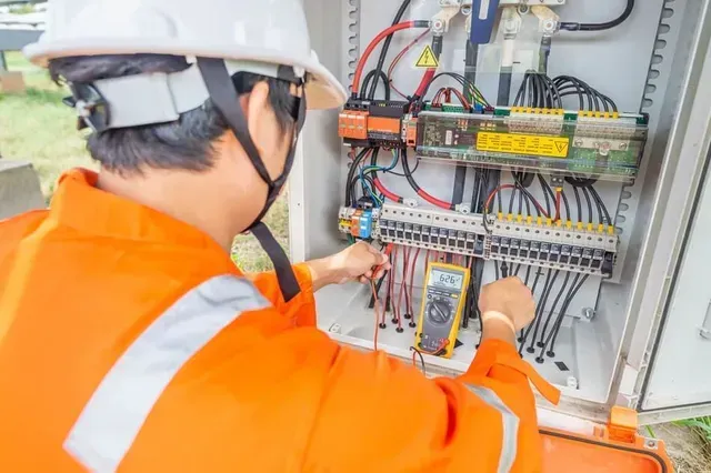 Un electricista con un mono naranja prueba el cableado en una caja eléctrica de metal con un multímetro.