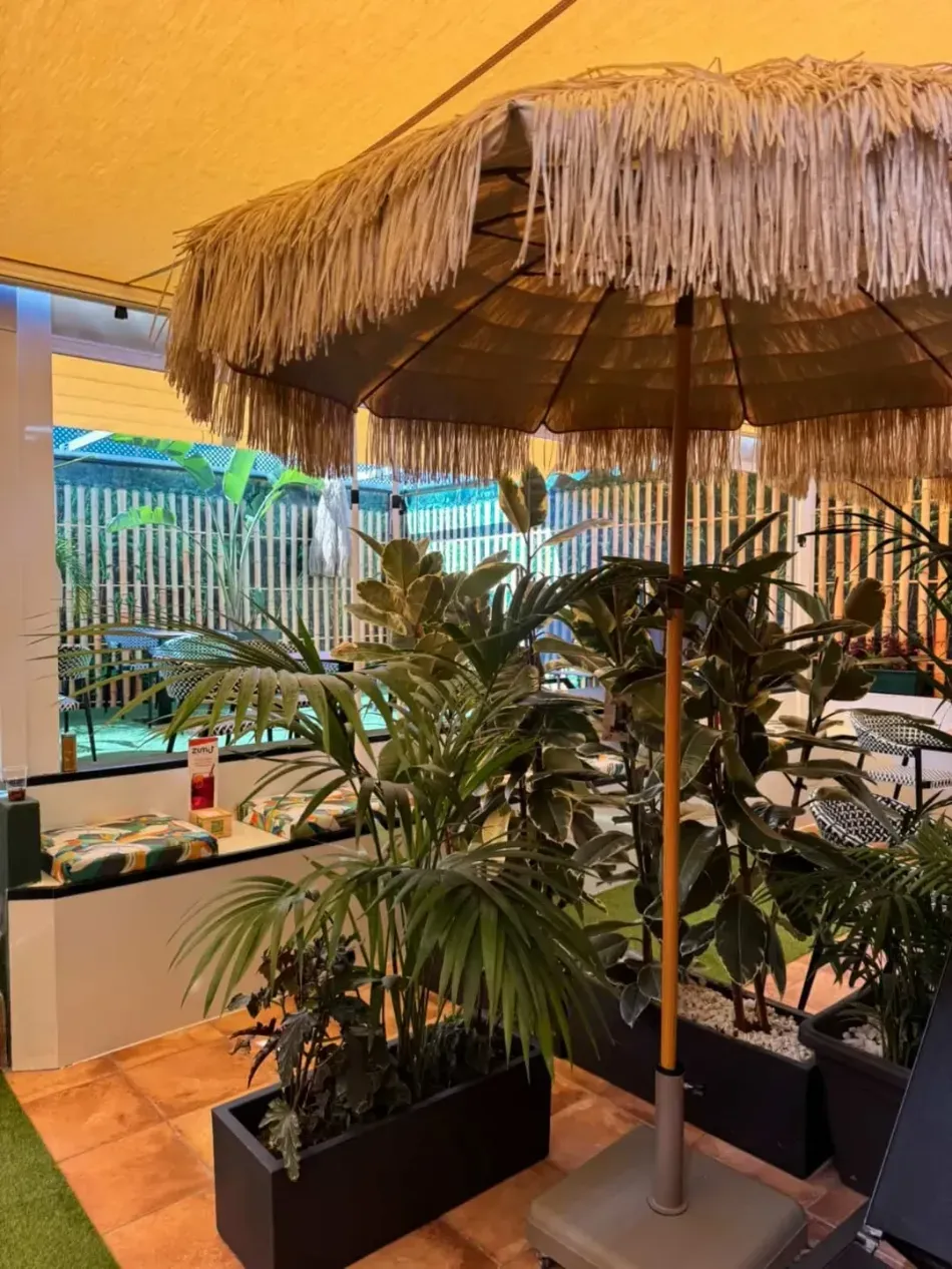 Patio interior con plantas tropicales en macetas bajo una sombrilla de paja y una cálida iluminación ambiental.