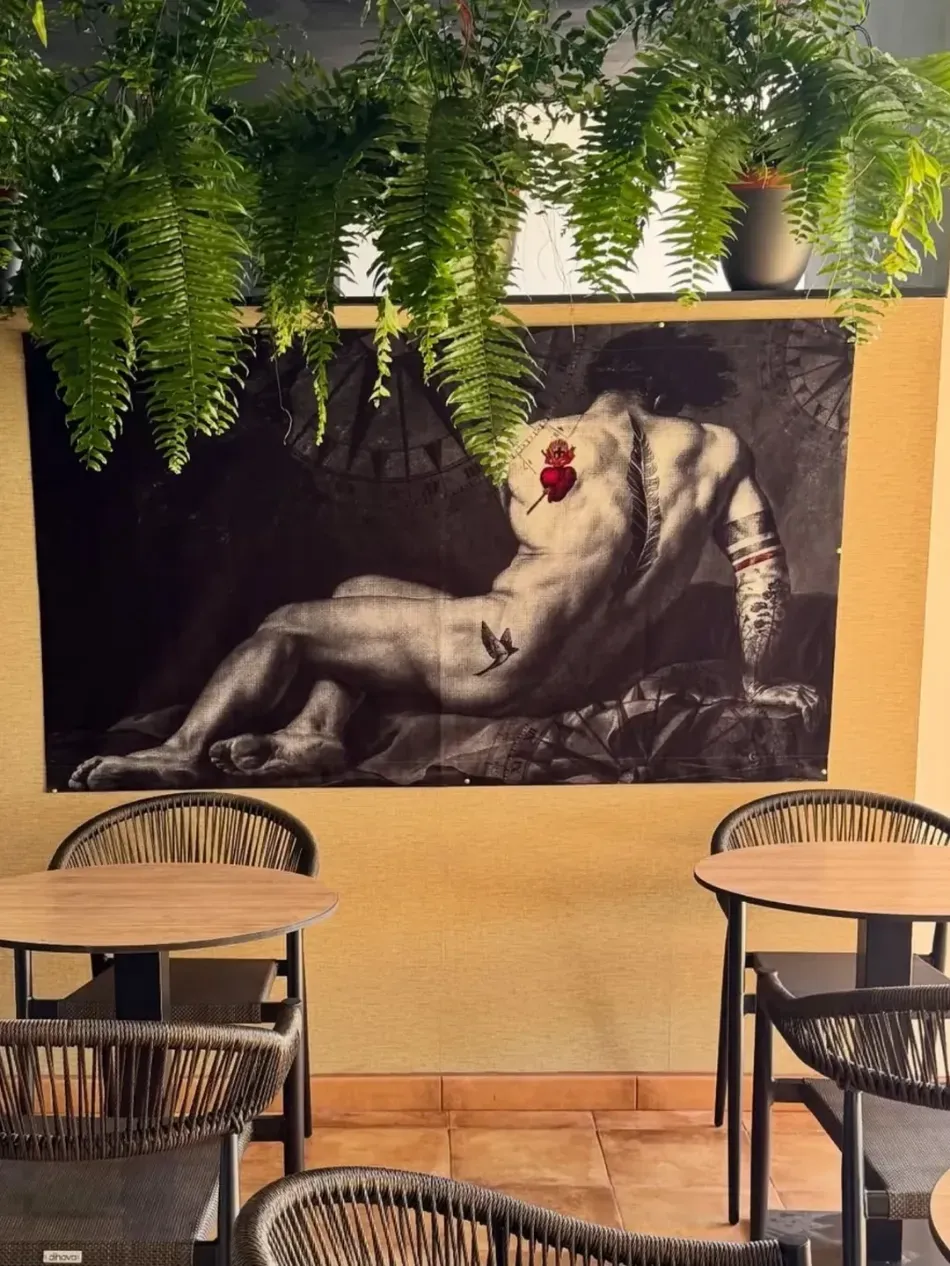 Pared de una cafetería con un gran mural en blanco y negro de una figura reclinada, plantas colgantes y mesas y sillas vacías.