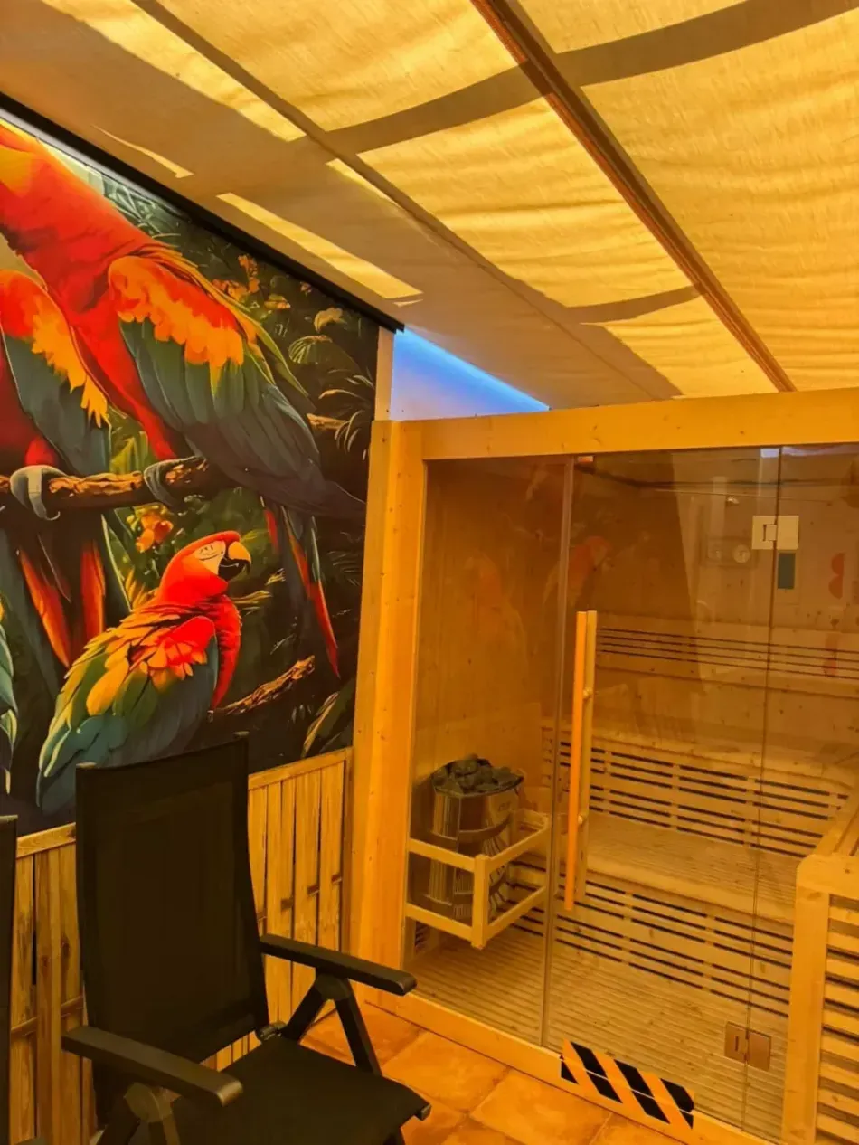 Mural tropical junto a una cabaña de sauna de madera con puerta de cristal y silla negra en un cálido espacio interior.
