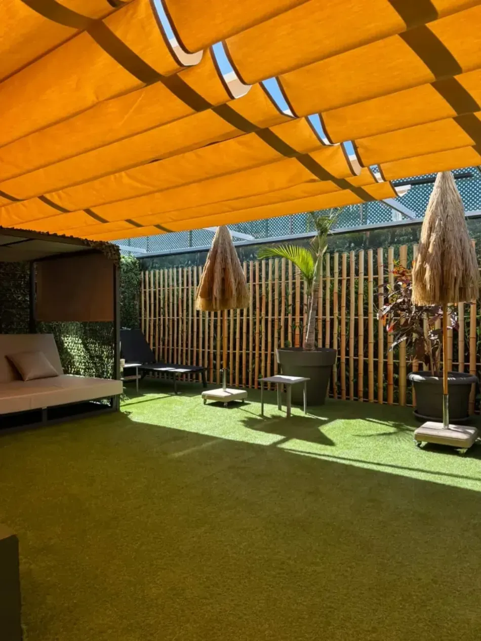 Patio exterior sombreado con toldo naranja, césped verde, tumbona, mesa y sombrillas de paja.
