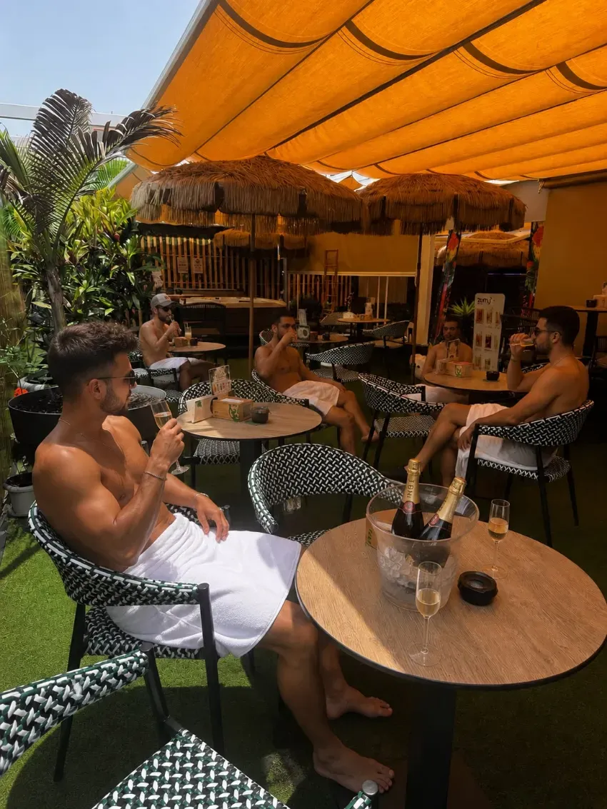 Gente relajándose en mesas de café al aire libre bajo un toldo naranja, con bebidas y vegetación cerca.