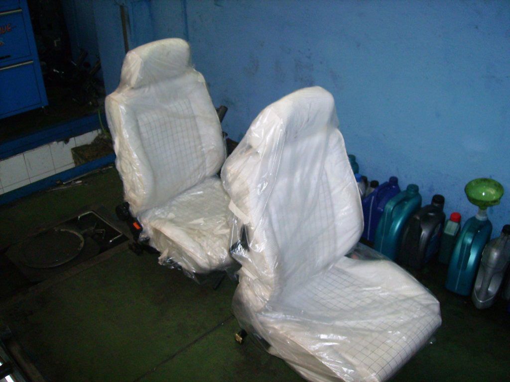Dos asientos de coche nuevos, blancos y cubiertos de plástico en un garaje.