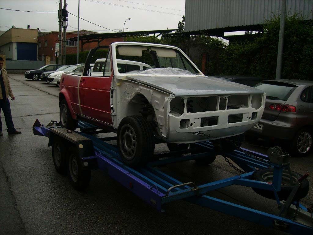 Volkswagen descapotable rojo y gris, parcialmente ensamblado, sobre un remolque azul, estacionado en una carretera mojada.