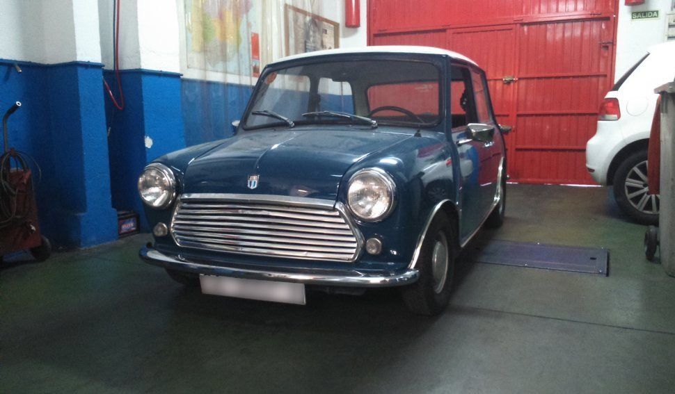 Mini Cooper clásico azul en un garaje con puerta roja y coche blanco parcialmente visible.