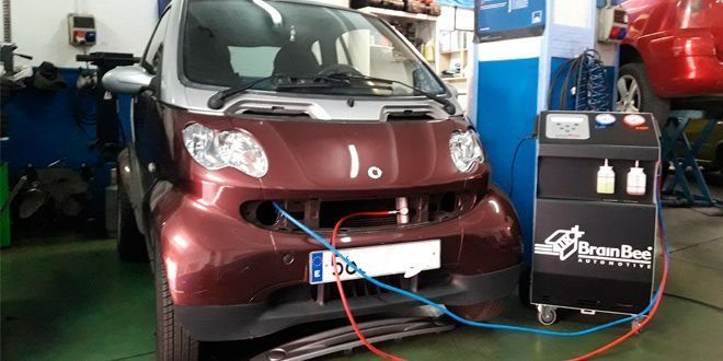 Un Smart color granate está siendo revisado, con mangueras azules y rojas conectadas al parachoques delantero. Una máquina está al lado del coche.