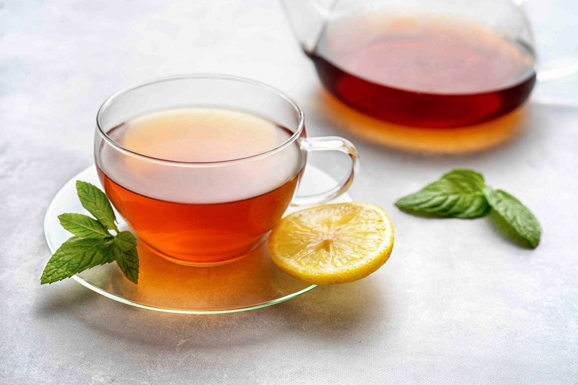 Eine Tasse Tee mit Zitrone und Minzblättern auf der Untertasse, im Hintergrund eine Teekanne.