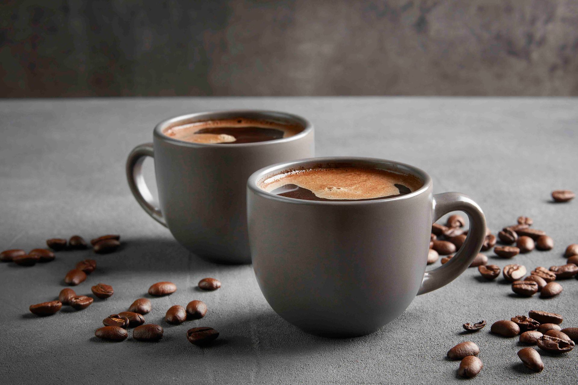 Zwei graue Kaffeetassen, gefüllt mit Kaffee, umgeben von Kaffeebohnen auf grauem Untergrund.