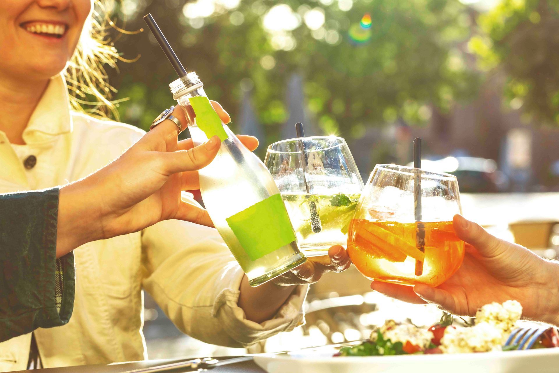 Menschen stoßen an einem sonnigen Tag im Freien mit Cocktails und einer Flasche Wein an.
