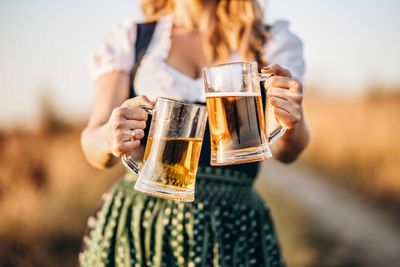 Eine Frau in traditioneller deutscher Tracht hält auf einem Feld zwei Bierkrüge hoch.