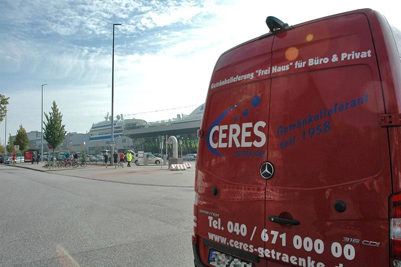 Ein roter CERES-Lieferwagen steht in einer Stadtstraße und verdeckt teilweise ein Gebäude und Passanten