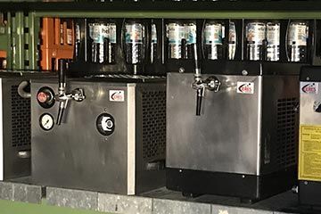 Zwei Bierfasskühler aus Edelstahl mit Zapfhähnen vor einer Barwand mit Biermarken.