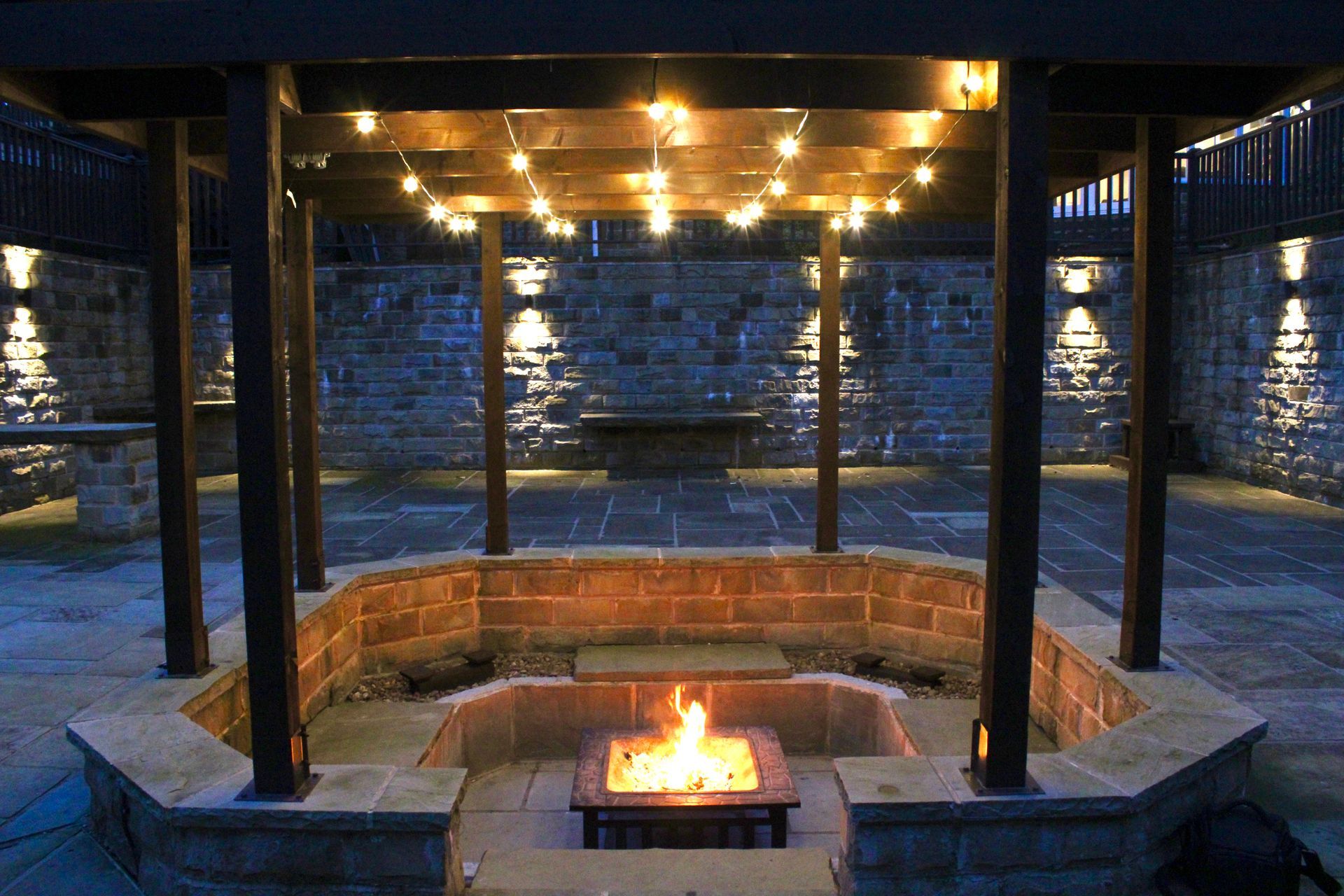 Nighttime outdoor patio with a fire pit, string lights, and stone walls.