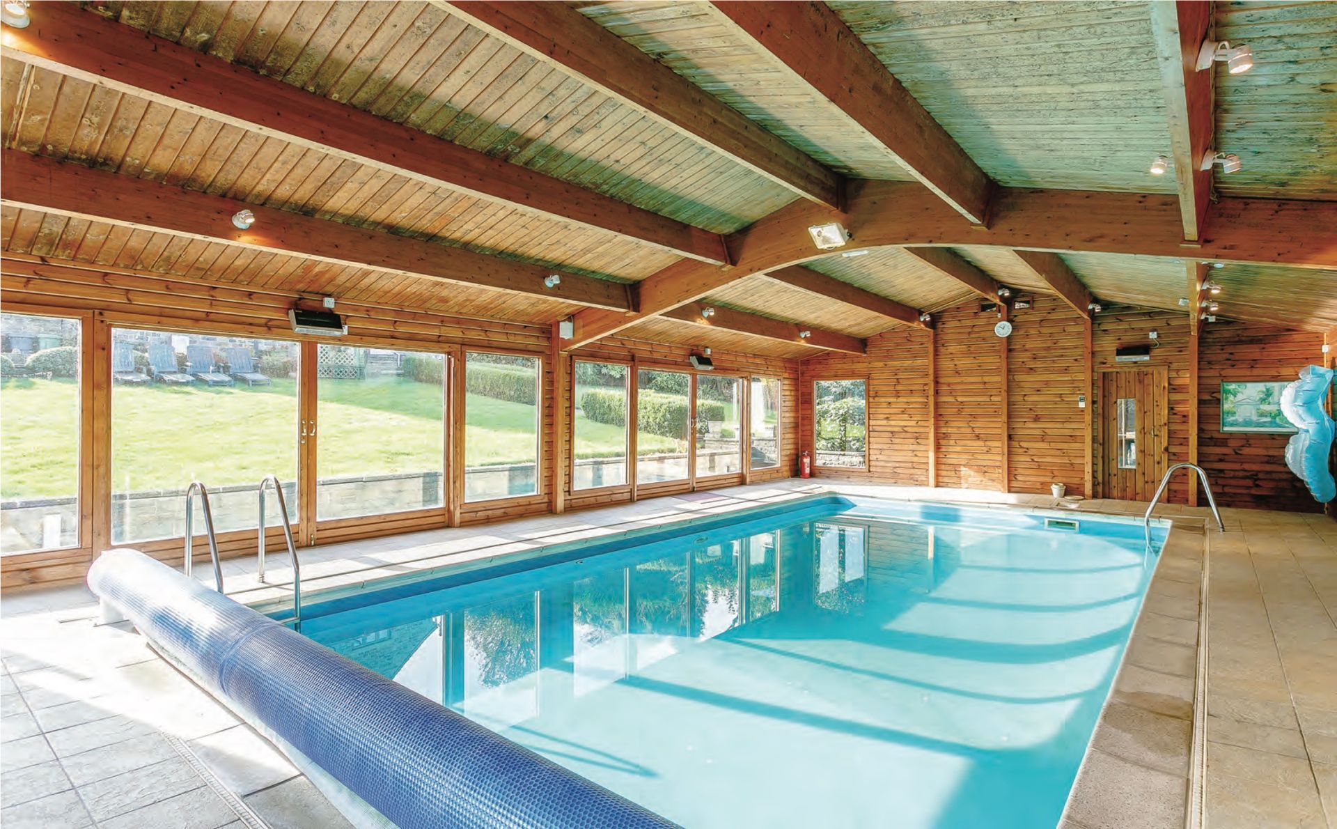 Indoor swimming pool with wood paneling and large windows overlooking a grassy landscape.