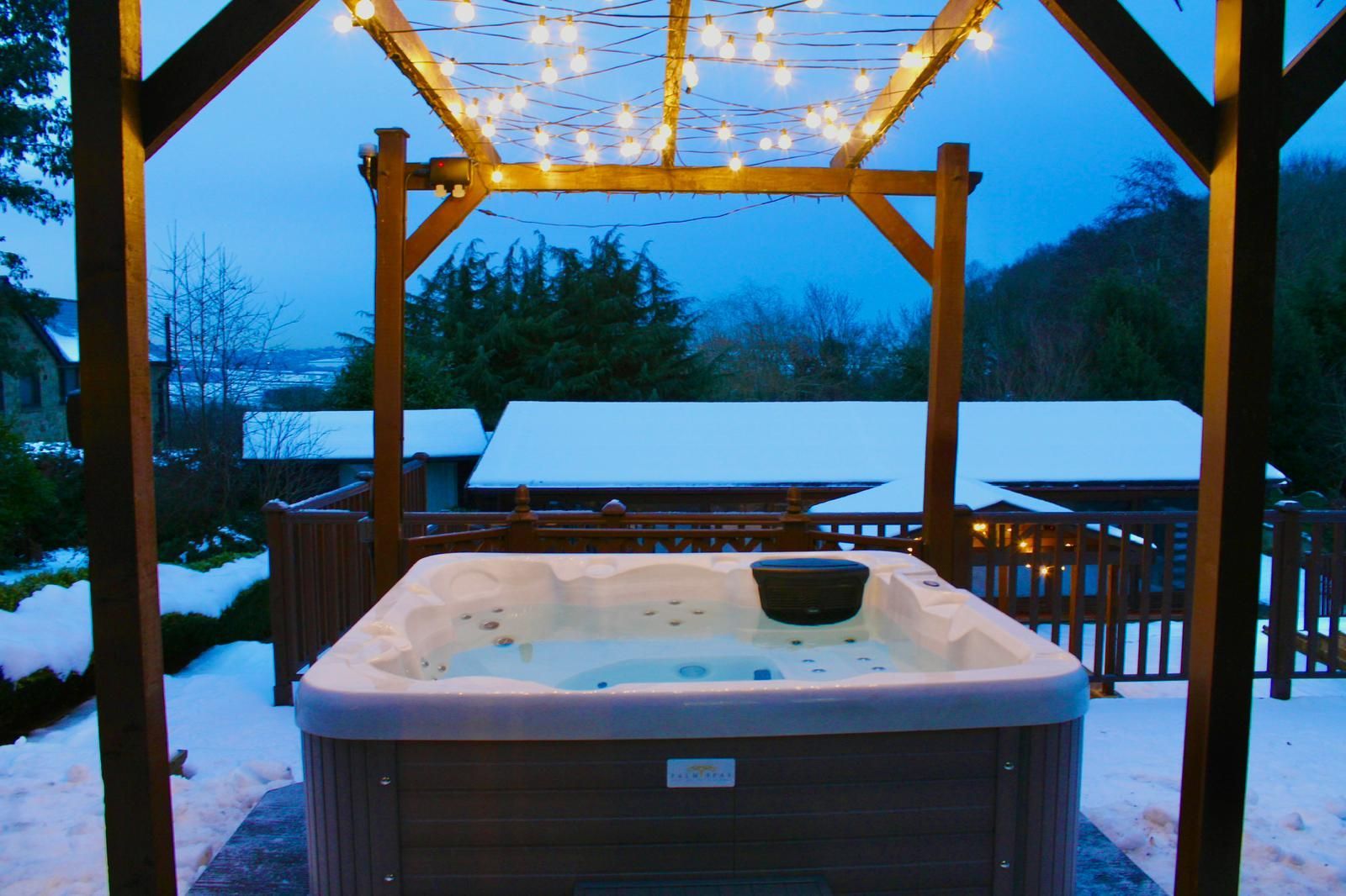 Hot tub under a wooden pergola with string lights, in a snowy outdoor setting.