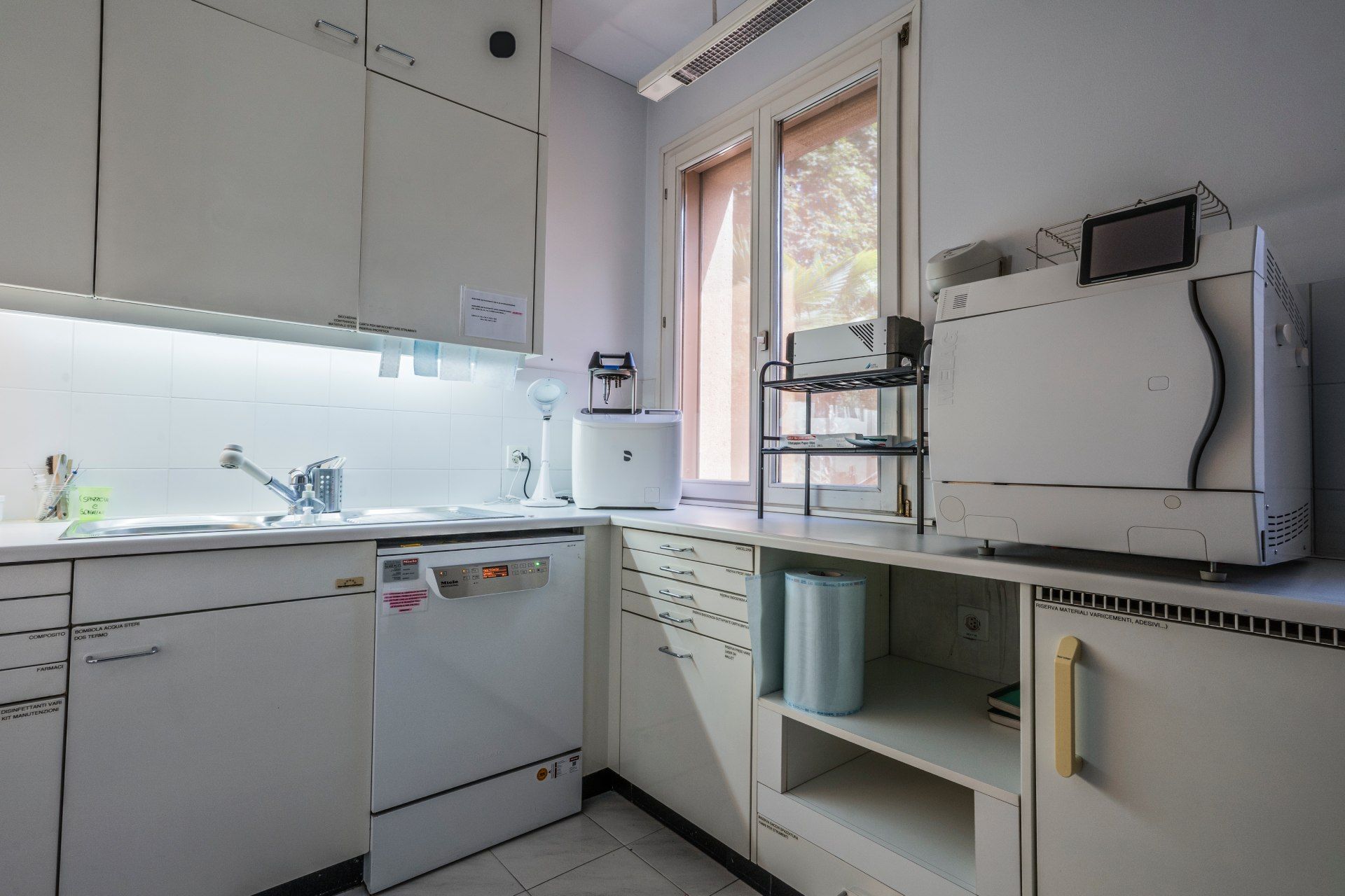 Interno bianco di un laboratorio medico con lavandino, armadietti, finestra e attrezzatura.