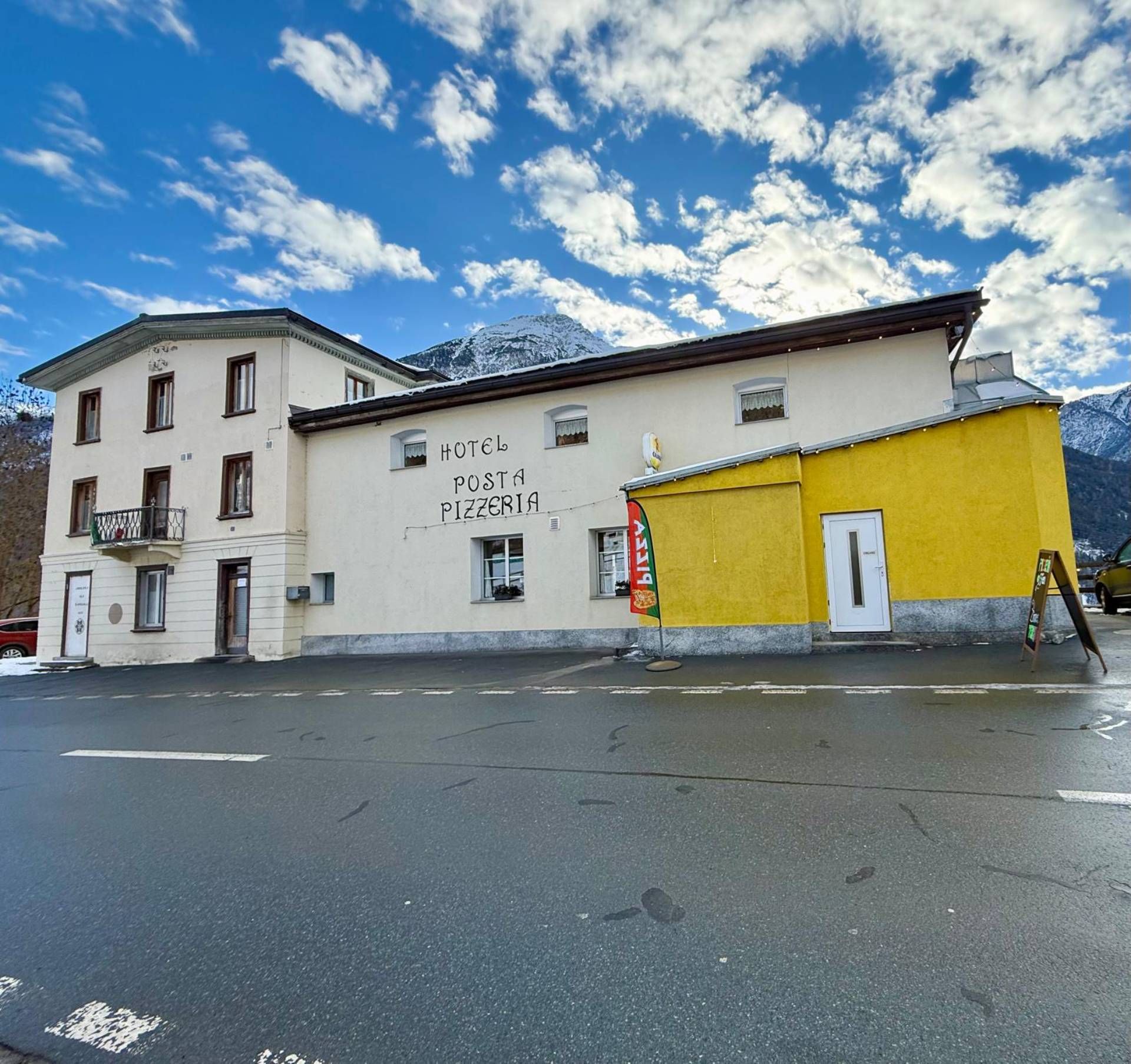 Hotel mit dem Schild „Hotel Plaza“, weisser Fassade, gelber Seite, Asphaltstrasse, im Hintergrund schneebedeckte Berge.
