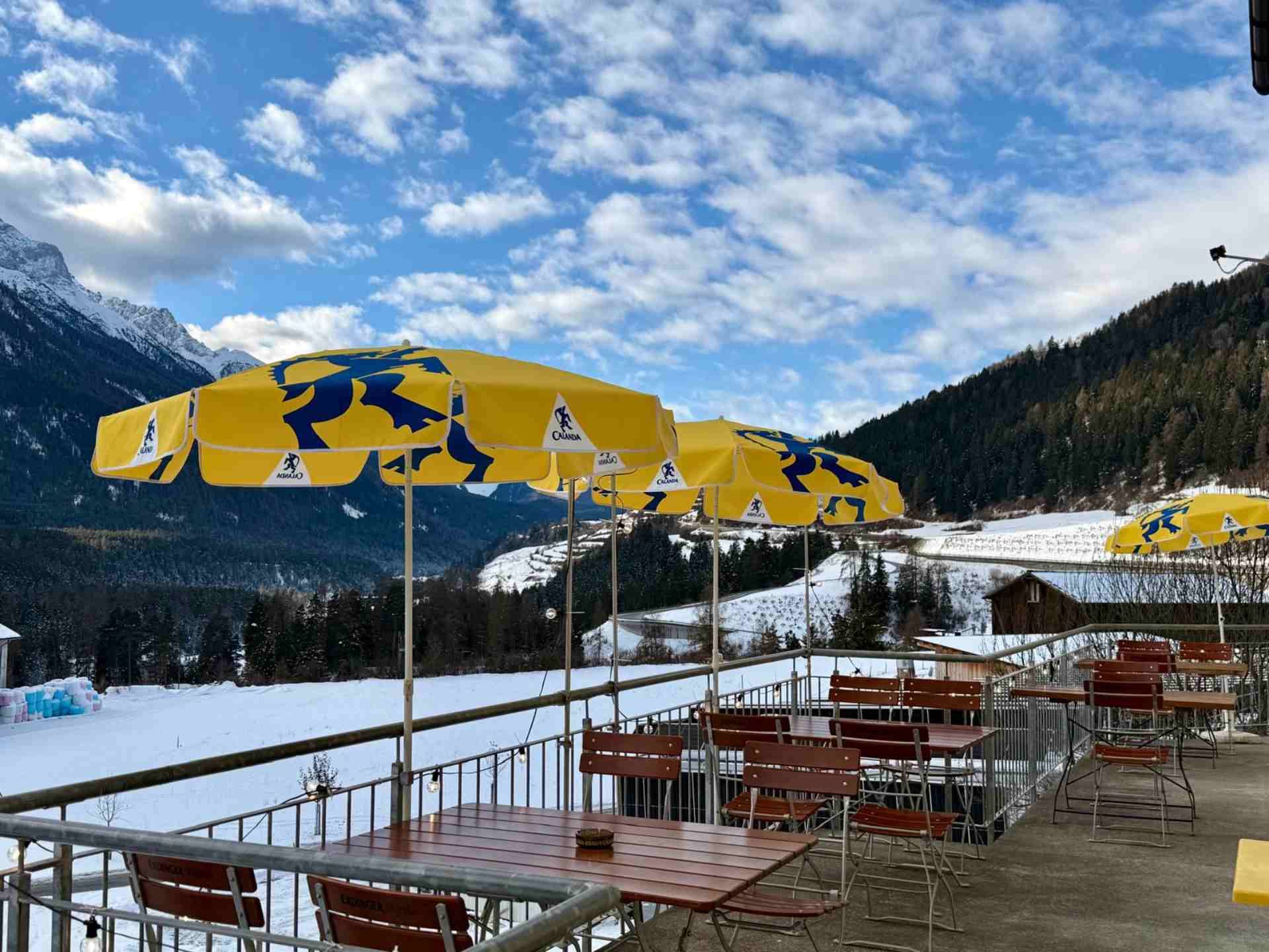Restaurant im Freien mit gelben Sonnenschirmen, schneebedecktem Boden und Bergkulisse unter bewölktem Himmel.