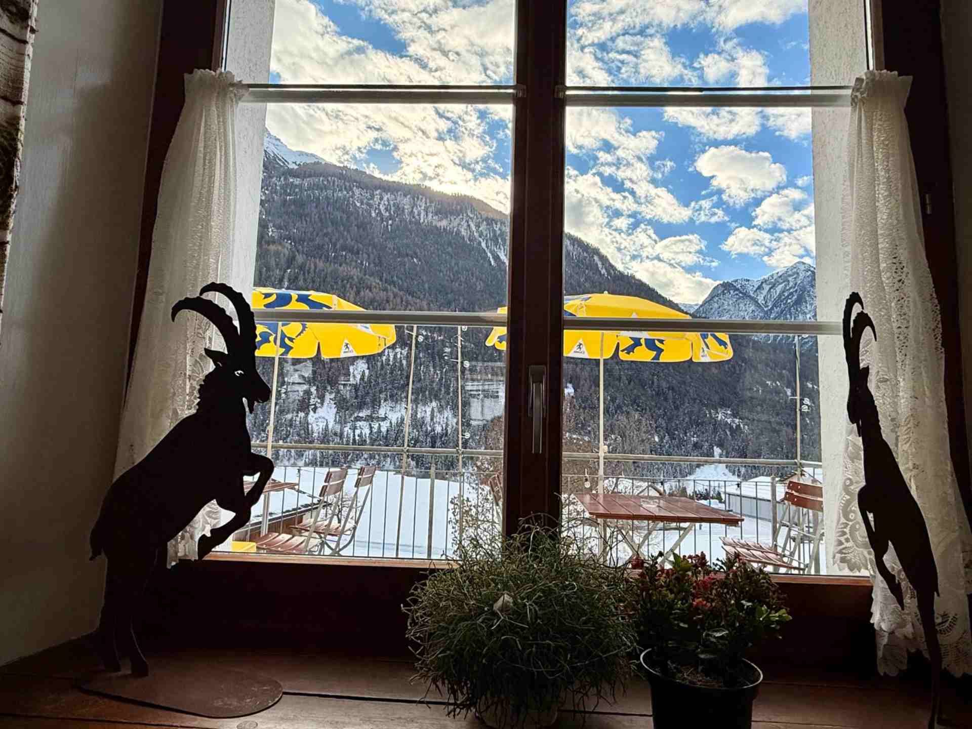 Blick aus dem Fenster: schneebedeckte Berge, blauer Himmel, zwei Ziegensilhouetten und ein Balkon mit gelben Sonnenschirmen.