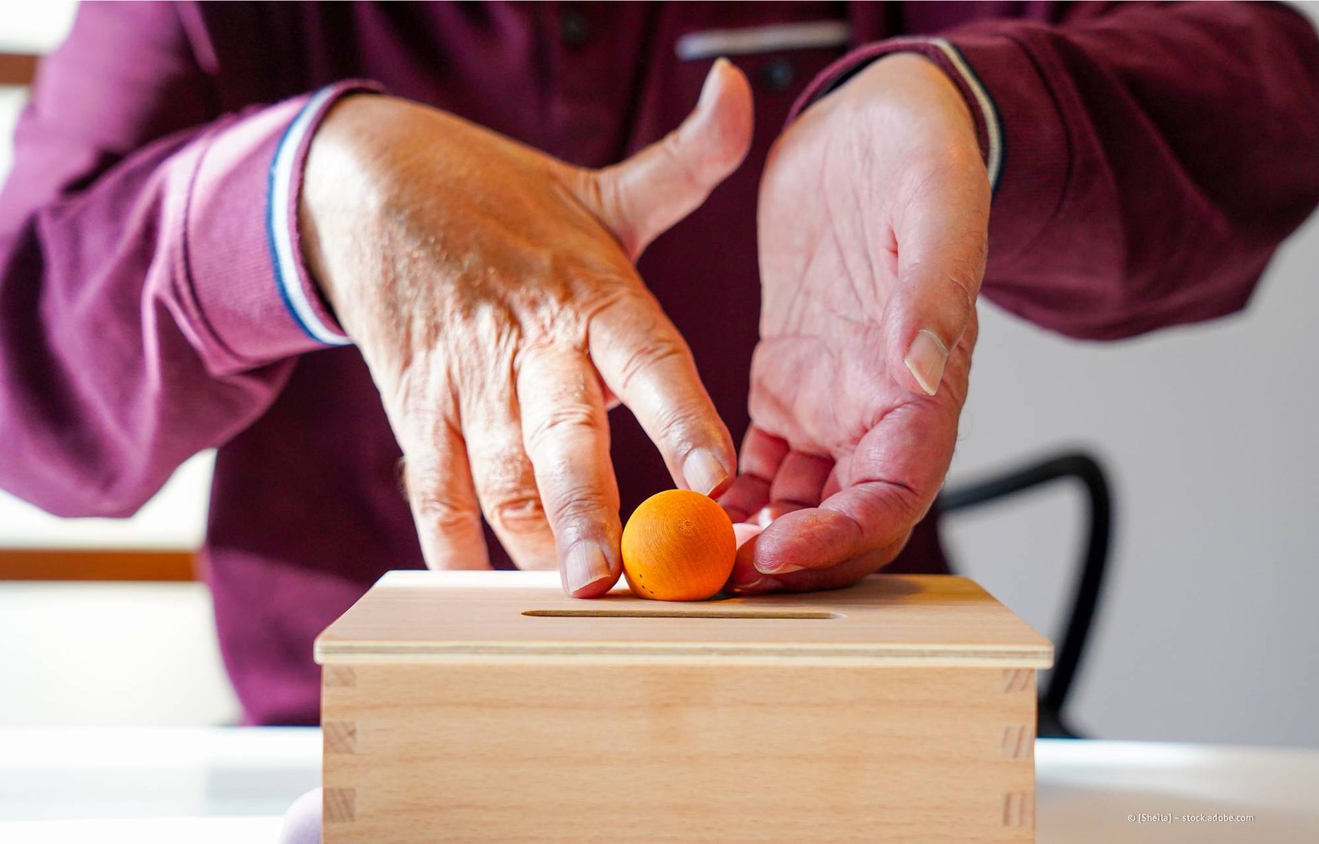 Hände greifen nach einem kleinen orangefarbenen Ball, der auf einer Holzkiste mit einem Schlitz liegt.