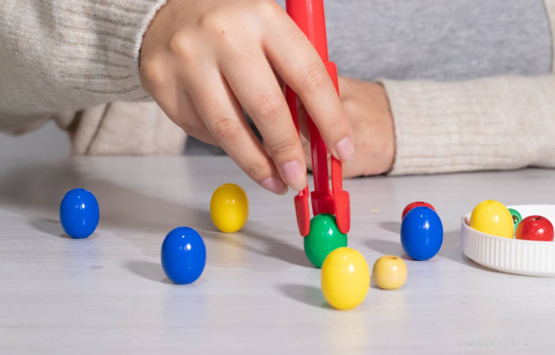 Eine Person nimmt mit einer roten Pinzette eine grüne Perle von einem Tisch mit verschiedenfarbigen Perlen.