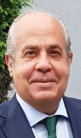 Hombre de tez clara, vestido con traje y corbata, sonriendo levemente, al aire libre.