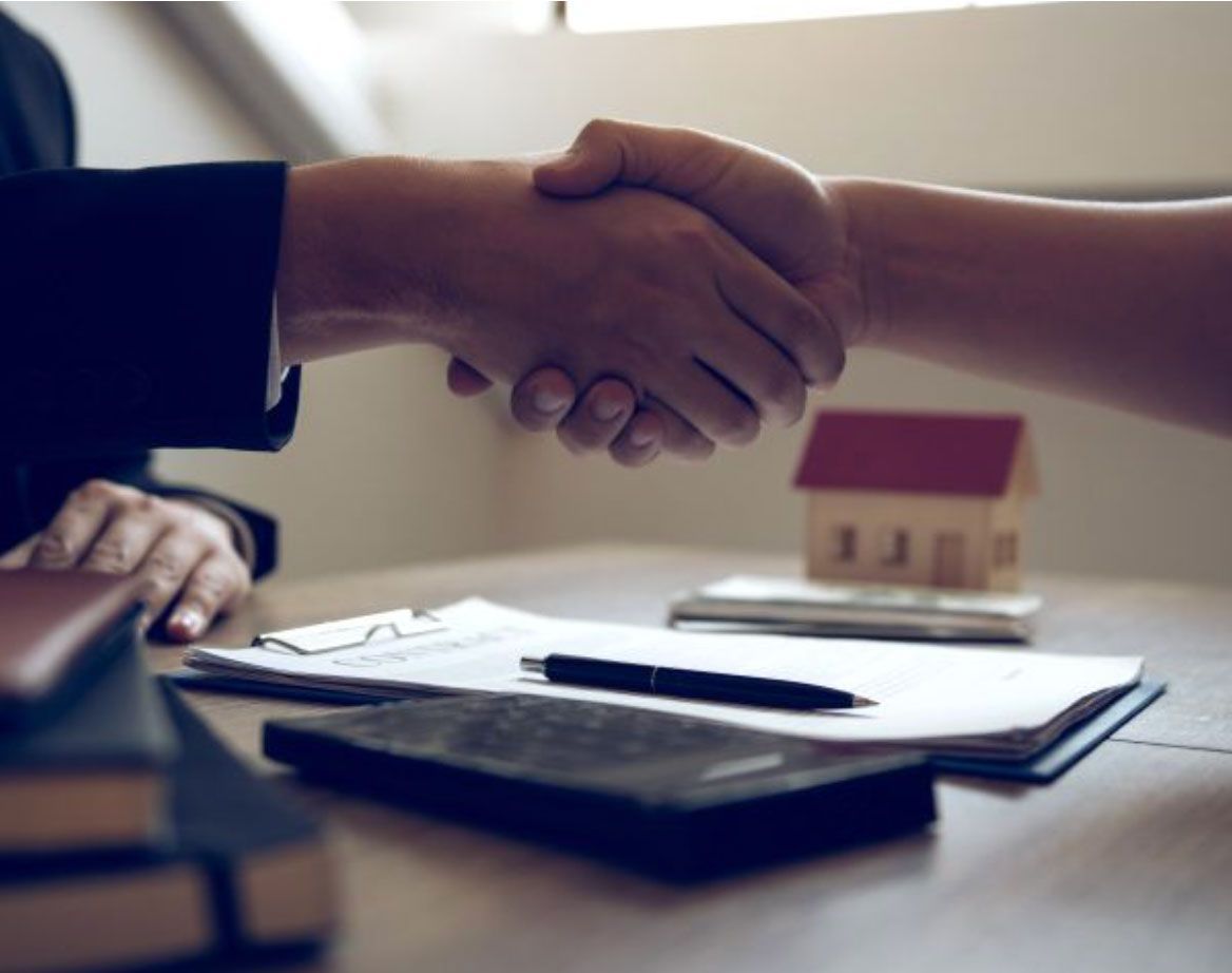 Dos personas se dan la mano sobre una mesa con una maqueta de casa, documentos y un bolígrafo, probablemente una operación inmobiliaria.