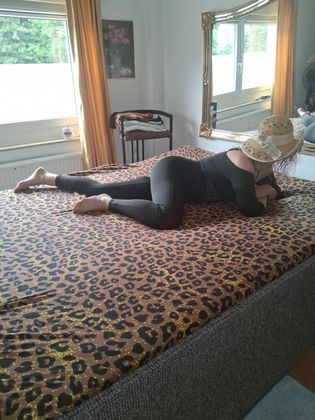 Eine Person liegt in einem hellen Schlafzimmer auf einem Bett mit Leopardenmuster. Sie trägt schwarze Kleidung und einen Hut.