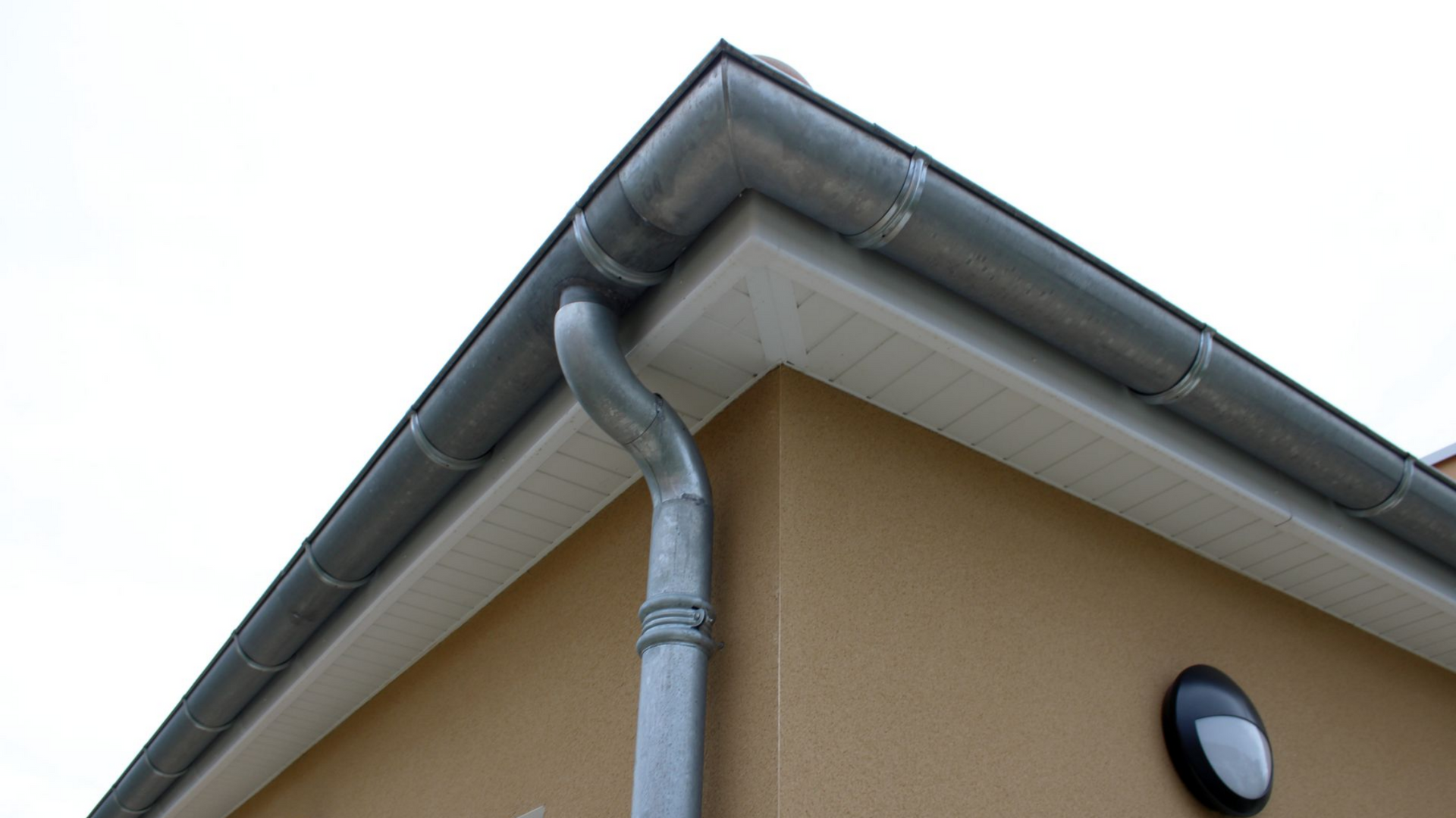 Gouttière grise à l'angle d'une maison beige avec dessous de toit blancs.