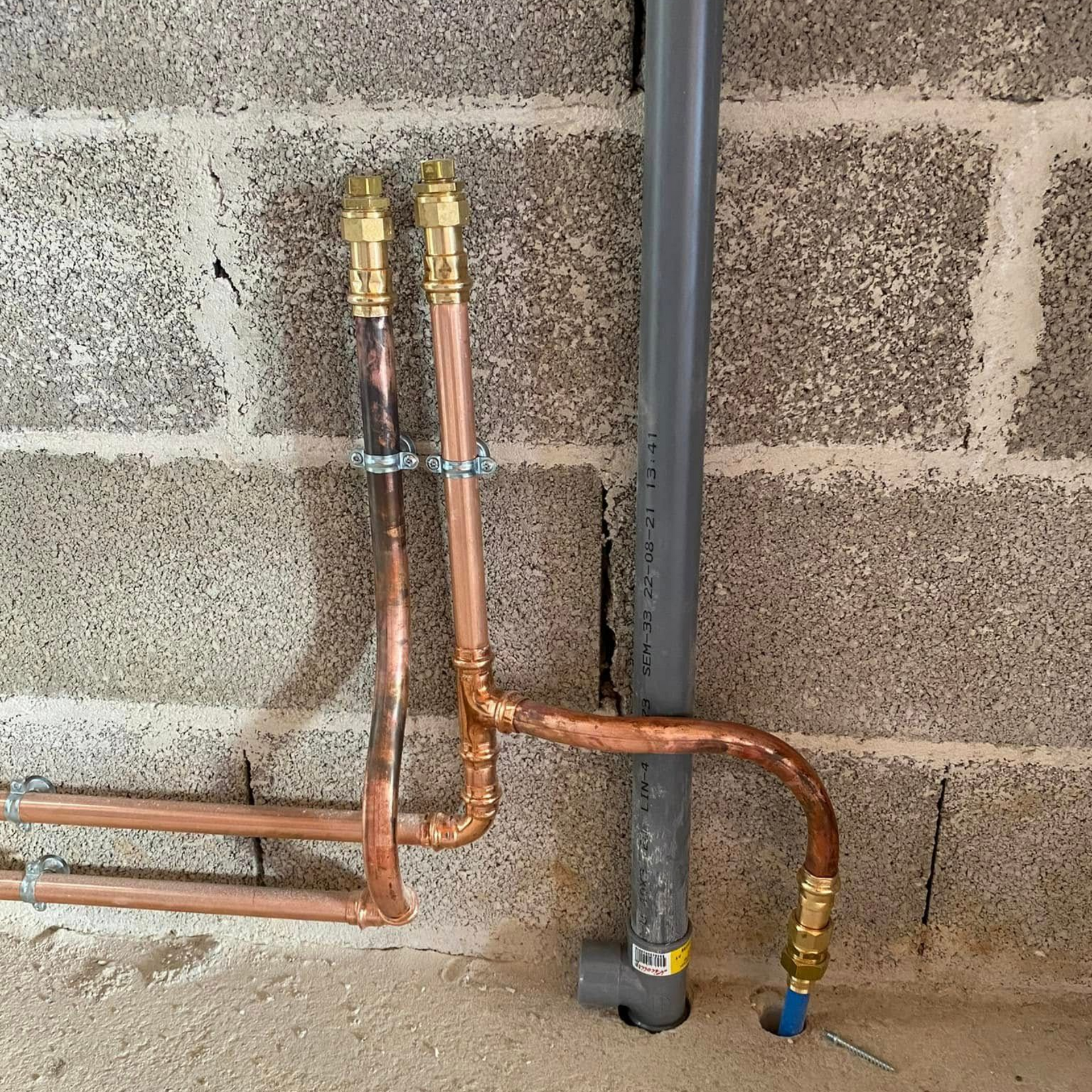 Des tuyaux d'eau en cuivre avec des raccords en laiton sont installés contre un mur en parpaings de béton gris, à côté d'un tuyau vertical en PVC gris