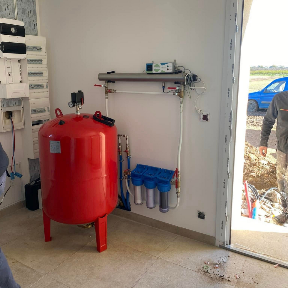 Un réservoir sous pression rouge relié à un système de filtration d'eau est fixé sur un mur blanc dans une pièce proche d'une porte ouverte sur l'extérieur