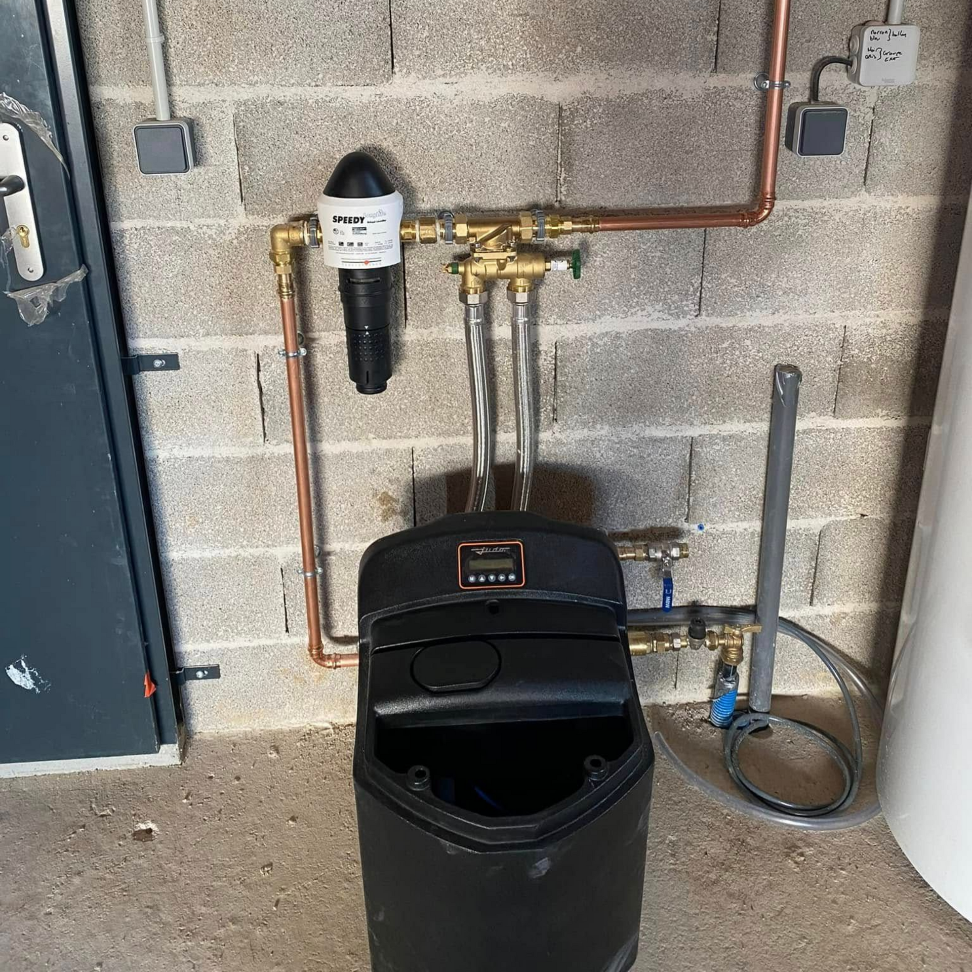 Système d'adoucisseur d'eau avec unité noire, filtre à sédiments et tuyauterie en cuivre, monté sur un mur en parpaings