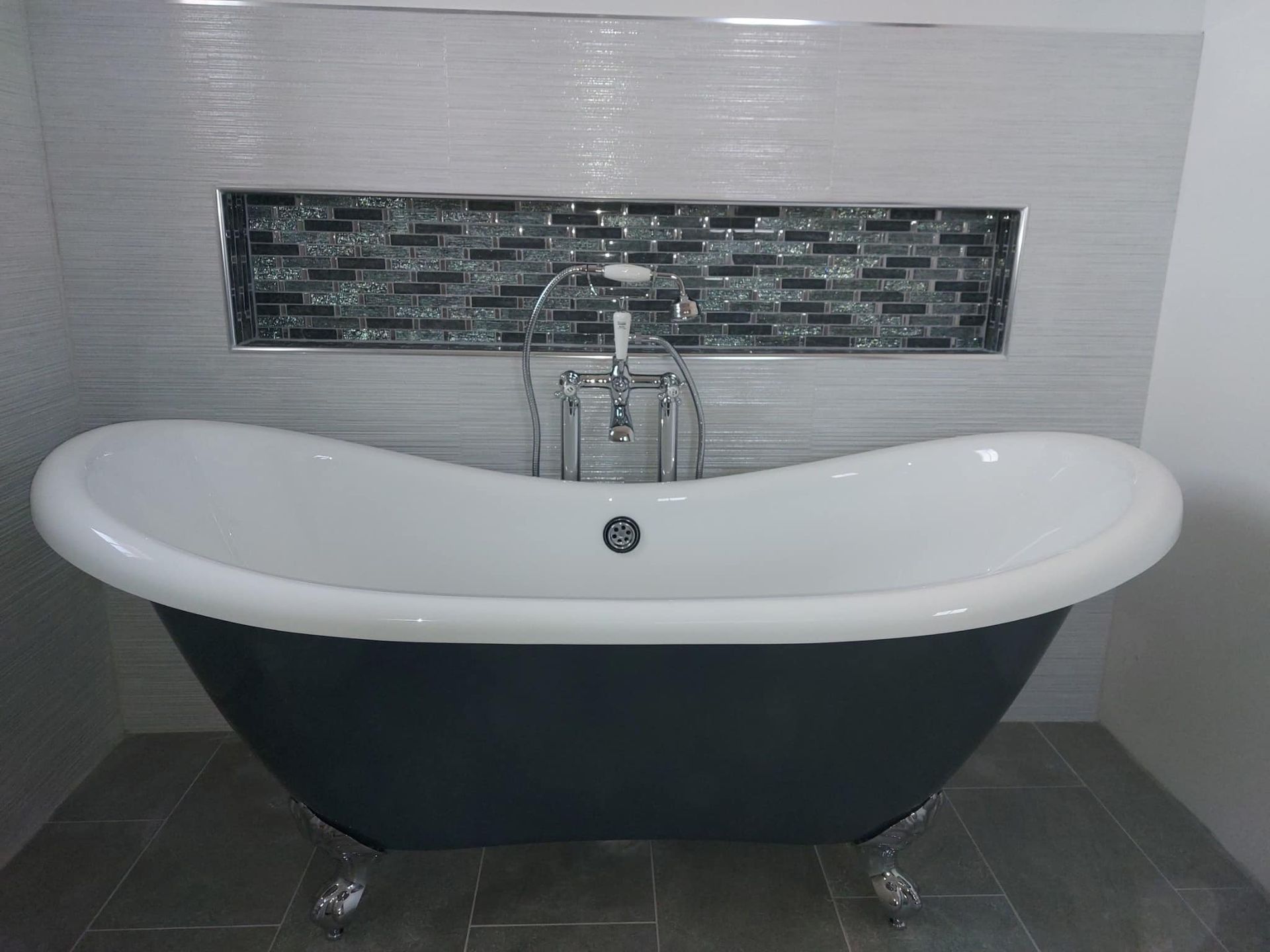 Une baignoire sur pieds gris foncé est adossée à un mur carrelé de gris clair et agrémenté d'une niche décorative en carreaux de verre foncé