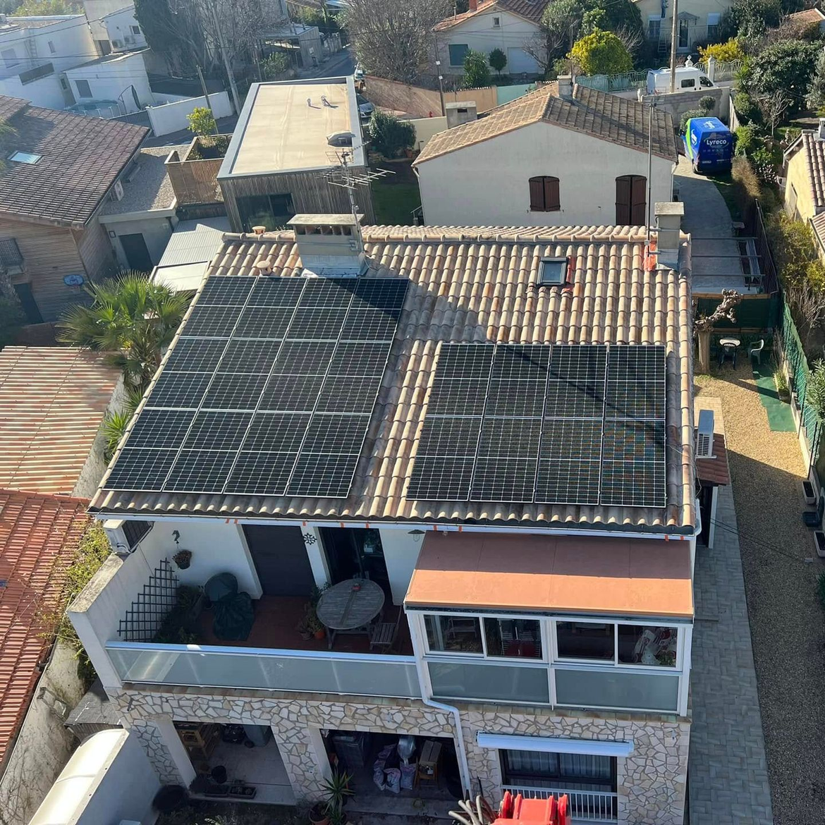 Vue aérienne d'une maison en pierre de deux étages avec des panneaux photovoltaïques sur son toit de tuiles, située dans un quartier résidentiel