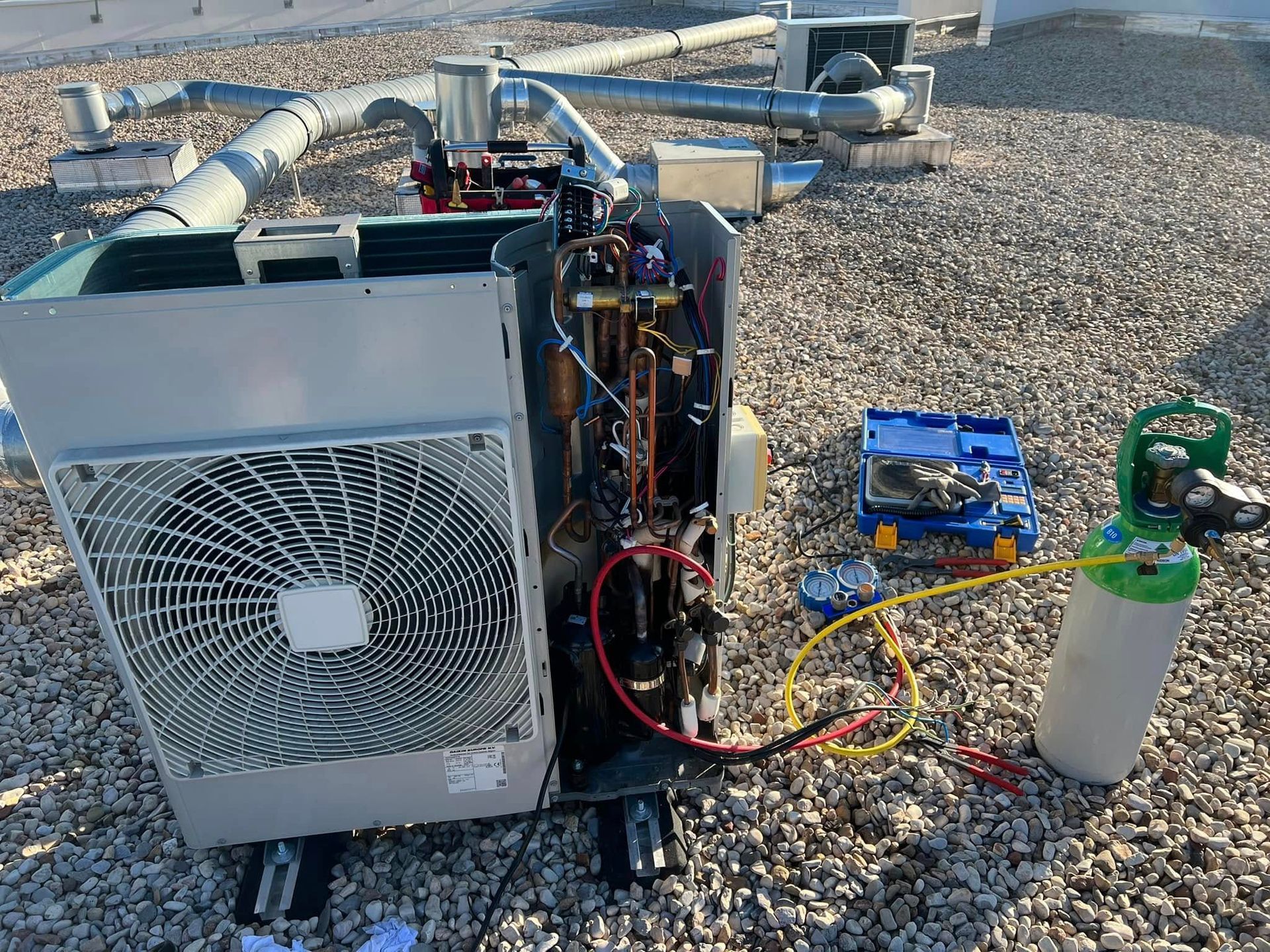 Un technicien effectue l'entretien d'une unité extérieure de climatisation sur un toit en gravier, avec un tuyau jaune relié à un réservoir de réfrigérant