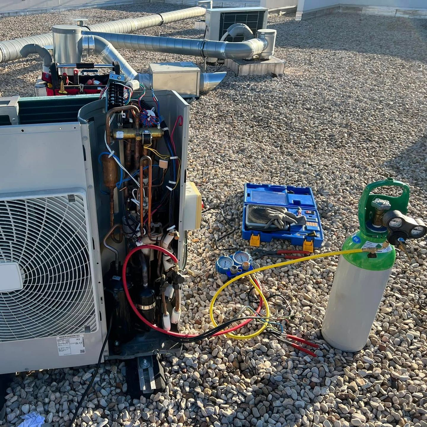 Unité de climatisation extérieure ouverte sur un toit en gravier, reliée à une bonbonne de réfrigérant par un tuyau de charge jaune