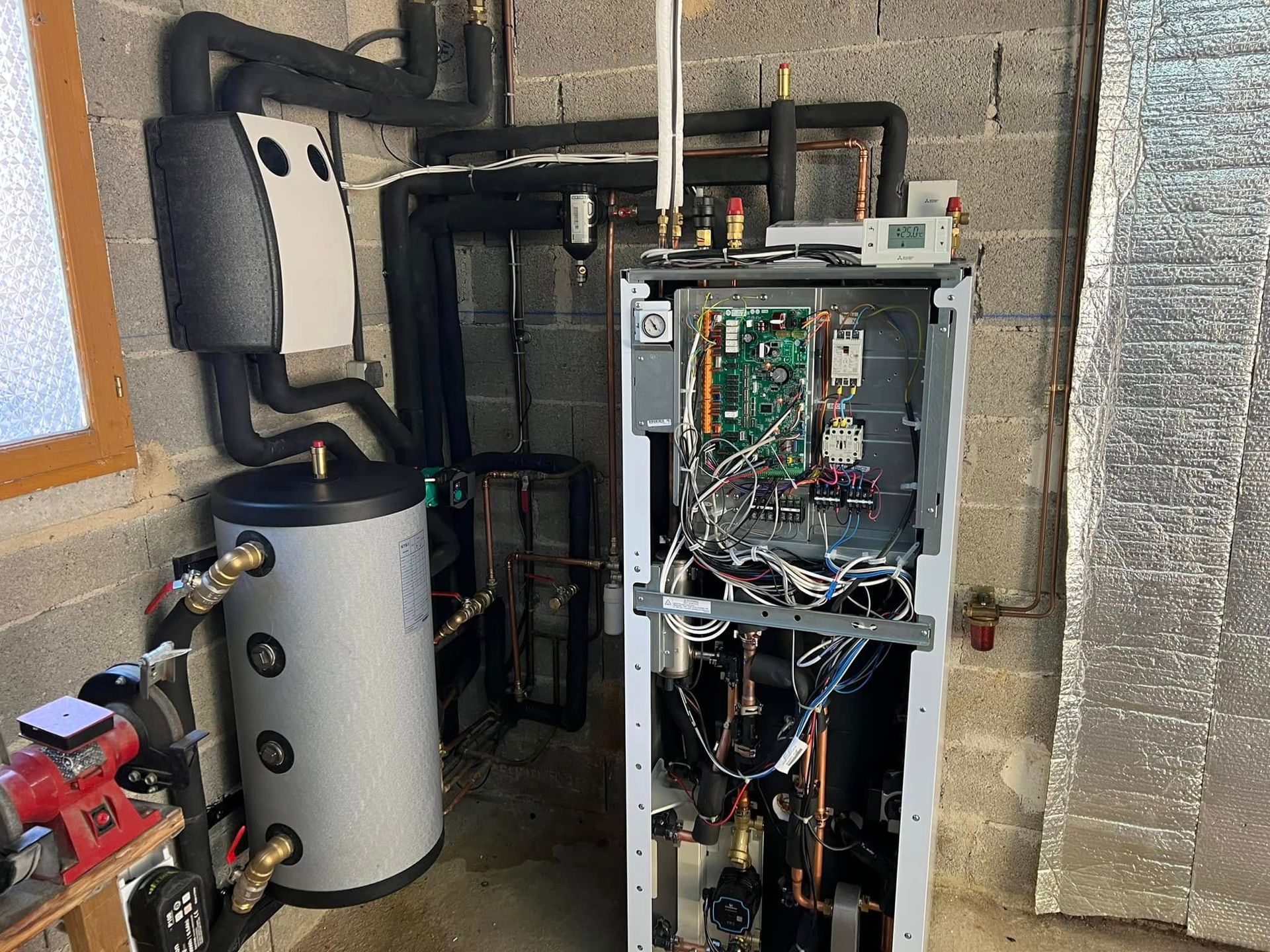 Système de chauffage dans une buanderie avec une chaudière haute, un réservoir d'eau cylindrique gris et des tuyaux isolés noirs