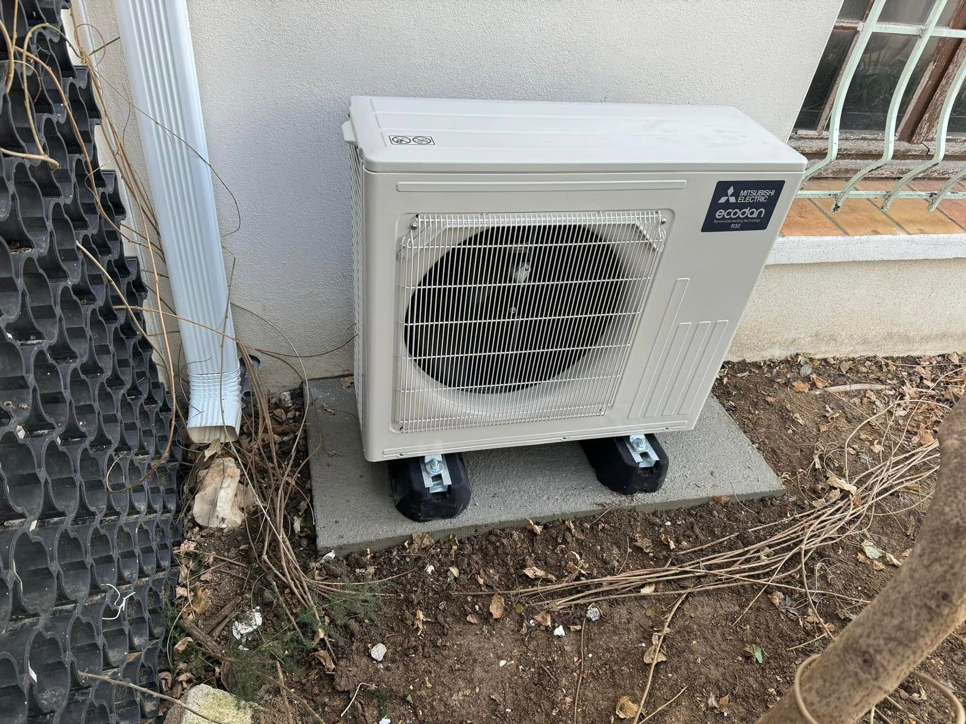 Un climatiseur extérieur Mitsubishi Electric de couleur claire est installé sur une dalle de béton près d'un mur et d'une clôture